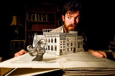 A bearded, fair-skinned man peering above a white paper creation of a building which rises up from the pages of a book. The building, and the forlorn figure of a woman holding a book to her chest, have been ornamented with ink on their paper foundations.