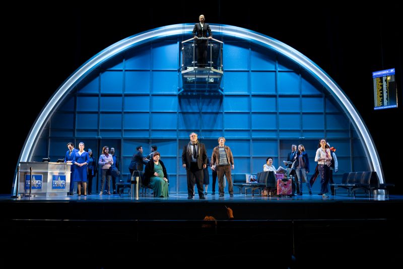 The cast of State Opera South Australia's 2025 production 'Flight'. A large group of performers stand on stage, which is designed to look like a nondescript airport. A final figure, The Controller, stands in a glass-walled balcony at the top of the photograph, as if monitoring everyone below.