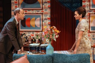 A man and a woman are standing and staring at each other over a kitchen table; a vase of flowers and a pair of bongo drums sit on the table. The wallpaper behind them is colourful and graphic.