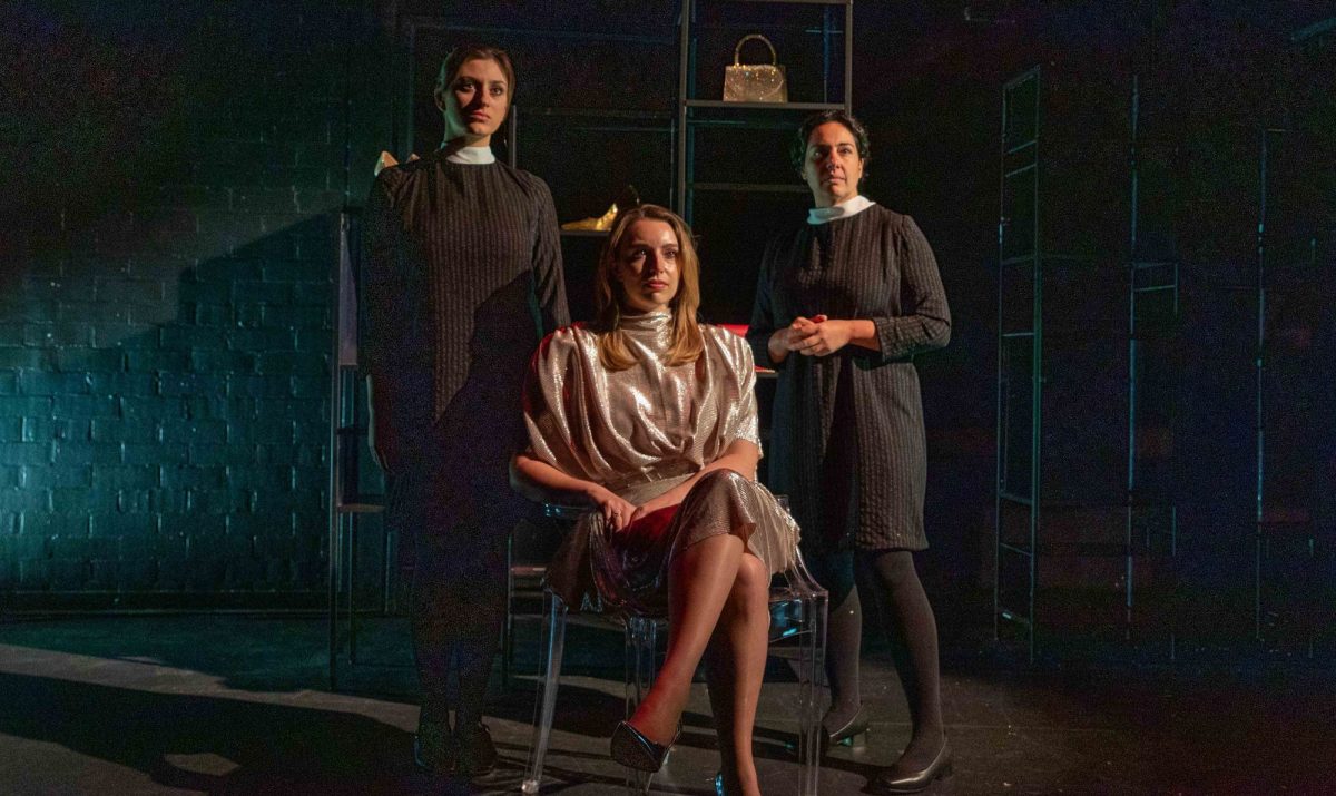 Two female-presenting actresses with dark hair in dark maid costumes stand stoically behind a blonde, female-presenting actor who wears a dress in a light-coloured fabric.