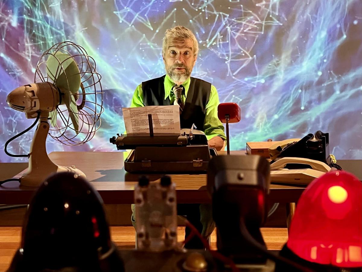 A man with grey beard and hair and wearing a lime shirt under a dark waistcoat is sitting at a table in front of a typewriter. Other random objects surround him including a fan.