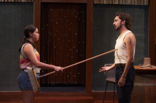 A young woman is poking the handle of a broom at a young man.
