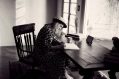 A woman with a hat sits perched over a desk, hand-writing into a journal on a wooden chair. The photo is in black and white. The woman is an author.
