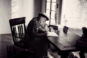 A woman with a hat sits perched over a desk, hand-writing into a journal on a wooden chair. The photo is in black and white. The woman is an author.