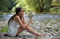 A fair-skinned woman sits barefoot on the pebbled bank of a river, engrossed in her book. She is fair skinned, has long dark hair and wears dark-framed glasses and a light, sleeveless, summery dress.