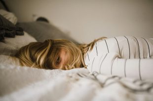 A woman with blonde hair has collapsed onto a bed. In this context of this article, it is demonstrating an artist with burnout.