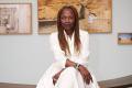 Koyo Kouoh: a dark skinned middle aged woman sitting in an art gallery with paintings behind her. She is wearing a white dress and has long braided hair. Venice Biennale 2026 curator.