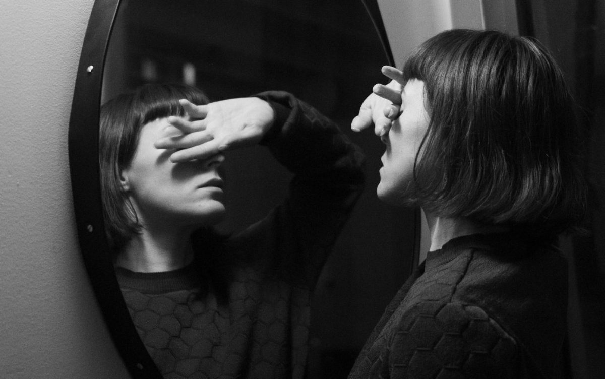 A black and white photo of a woman facing a round mirror with one hand covering her face.