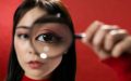 A figure holding a magnifying glass up to her eye, wearing pink eyeshadow and cat eyeliner. She is against a red background. Arts news.
