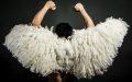 A performer wearing feathery wings with their back turned to the camera, holding both arms up in a powerful posture.