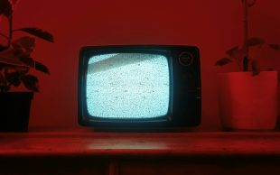 A photo of an old-school box television displaying noise. There are two pot plants on both sides of the tv, and the domestic scene is covered in a red tone.