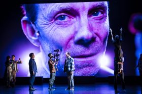 Theatre makers stand on stage as a close up of a mans face fills the theatre backdrop in a purple, blue hue.