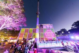 A dynamic and colourful photograph of Liverpool Powerhouse (formerly known as Casula Performing Arts Centre) at night. Crowds gather outside the venue; the building itself, including its tall smoke stack, is lit up with pink, yellow and green-toned projections.