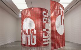 A floor to ceiling installation by Rose Nolan inside a gallery space. The installation comprises thousands of red and white hessian cloths, organised in a spiral formation with text that corresponds to the artwork title.