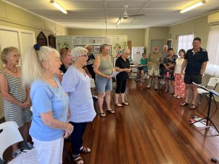 Members of the community choir rehearse for 'The Palace'. an artistic commemoration of the 25th anniversary of the Childers, Qld Backpackers' fire of 2000. Pictured are a group of 13 people, ranging in age from seniors to people in their 30s, rehearsing a song in a country hall meeting room.