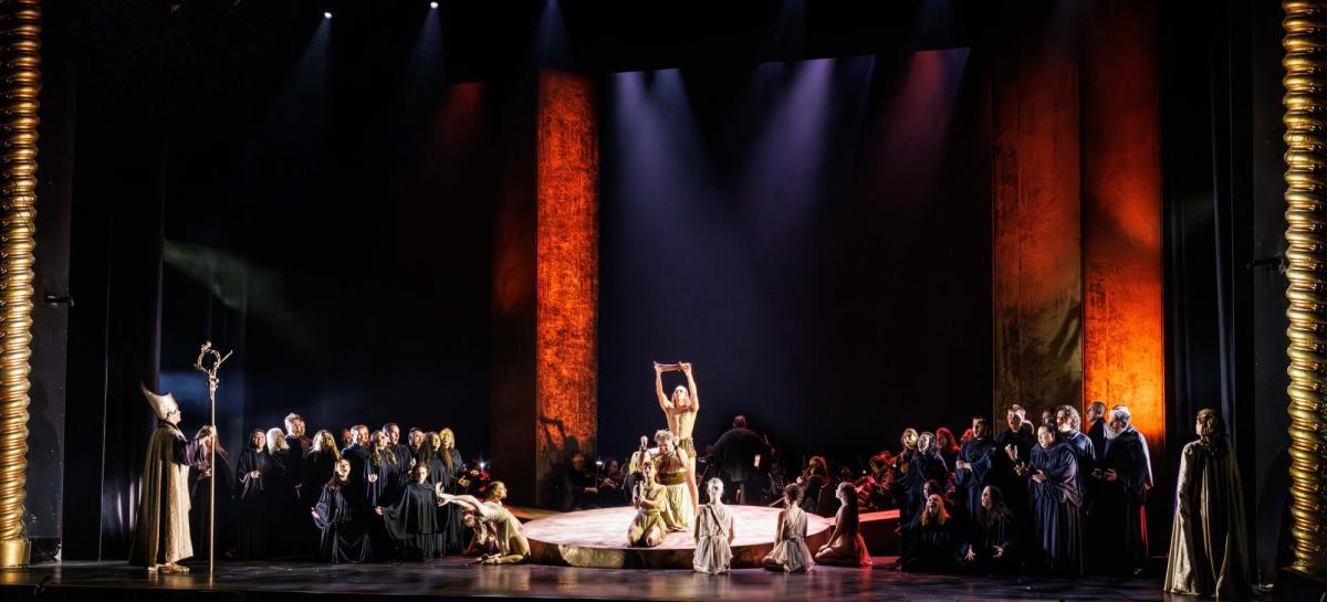 Large stage scene of 'Samson & Delilah'. In the centre are figures wearing gold costumes. They are surrounded by cast in black.
