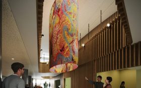 Digital render of a large oval cylinder tapestry with dots and curves hanging from the ceiling of the second floor and reaching down to the first floor. There are wood panelling inside the architectural foyer with people looking at the tapestry.