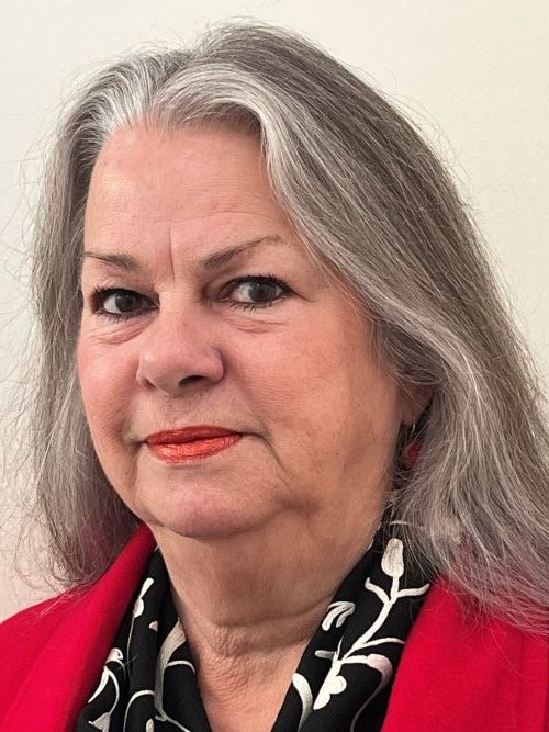 A fair skinned, fgrey-haired woman gazes into the camera. She is wearing a red coat over a black collared top pattered with a white floral design.