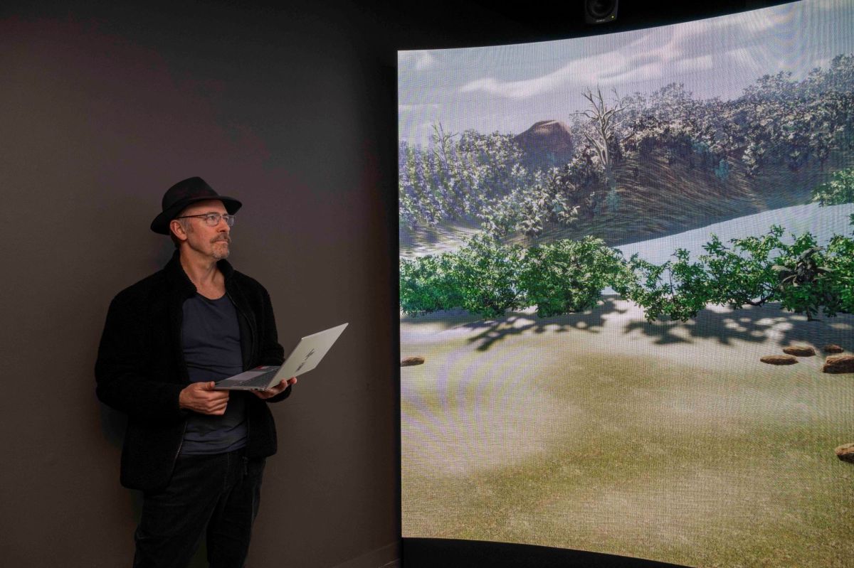 A older man wearing a black hat and black jacket holding a laptop and looking at a concave screen depicting a landscape with green shrubs.