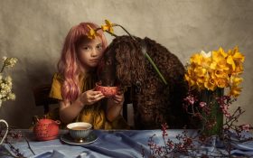 An artistic photo portrait of a young girl with long red hair feeding a long-haired dog next to her from a little orange mug. They are seated at a table with indigo blue table cloth and a yellow flower arrangement to the right.