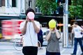 Two young people in a busy street wearing headphones, hold up a yellow and pink ball, blocking their faces.