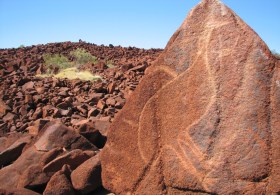Murujuga rock art: a landscape photograph of ochre red earth rocks, one of which has a line drawing/ engraving of an emu on it.