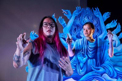 Joe Paradise Lui, a Chinese-Australian man with long red-streaked back hair and wearing glasses, stands with his hands raised in front of a representation of Guanyin, the Chinese bodhisattva or goddess of Mercy and Compassion, who is played by the Chinese-Australian actor and theatre-maker Merlynn Tong. She has short black hair and sits with her hands raised, in a blue prop representing Guanyin and her many arms and hands.