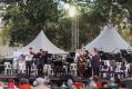 A musical recital in Townsville's Queen's Garden at the Australian Festival of Chamber Music 2024. The photo shows seven musicians playing a range of instruments including string and brass, on a low outdoor stage. under trees in front of a seated audience.