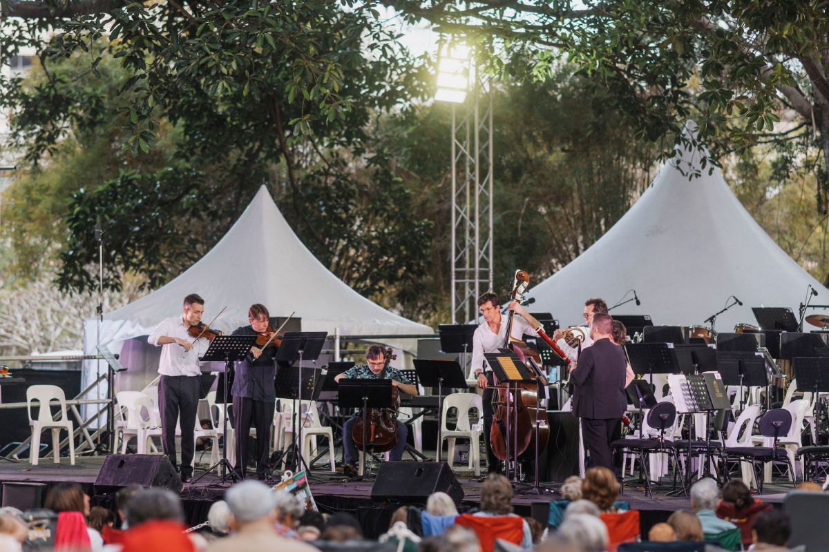 A musical recital in Townsville's Queen's Garden at the Australian Festival of Chamber Music 2024. The photo shows seven musicians playing a range of instruments including string and brass, on a low outdoor stage. under trees in front of a seated audience.