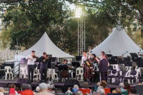 A musical recital in Townsville's Queen's Garden at the Australian Festival of Chamber Music 2024. The photo shows seven musicians playing a range of instruments including string and brass, on a low outdoor stage. under trees in front of a seated audience.