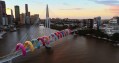 13 brightly coloured inflatable loops arch over a Brisbane Bridge. The photo is taken from above, showing the muddy river beneath and some of the city skyline.