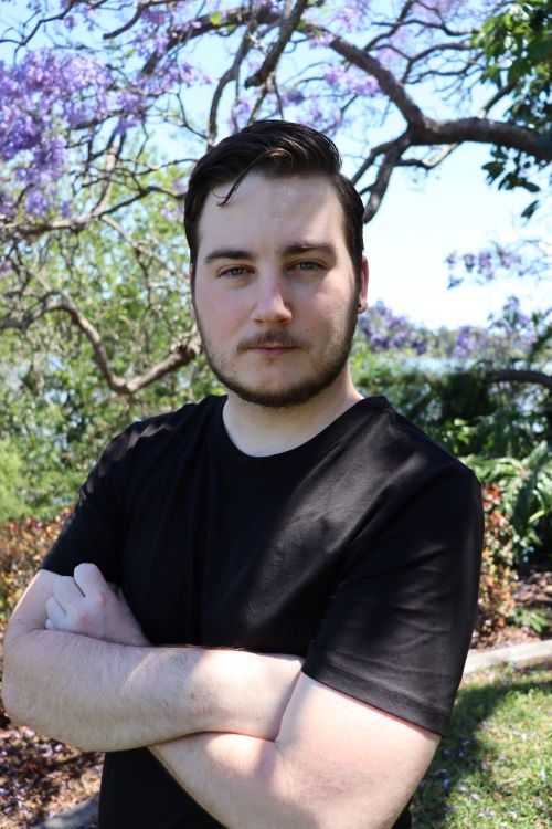 An Aboriginal man with light skin stares into the camera. He has black hair, parted on the left and swept over to the right; a single lock of hair falls over his forehead. He also has a short, closely trimmed black beard and moustache. He wears a black t-shirt and stands with his arms folded. Foliage is vible behind him.