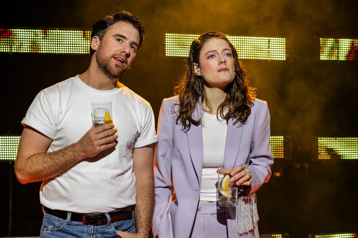 A man in a white T shirt and jeans and a woman in a lilac jacket stand on stage with drinks