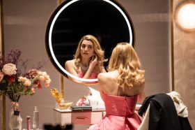 A glamorous woman in a pink silky gown sits in front of a mirror putting her earrings in. Aphrodite.