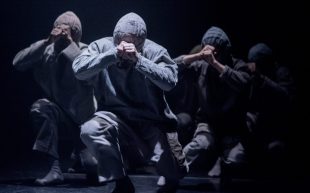 A close-up short of three dangers in the spotlighting squatting low and walking with their hands in a fist held up to their heads, covering their face.