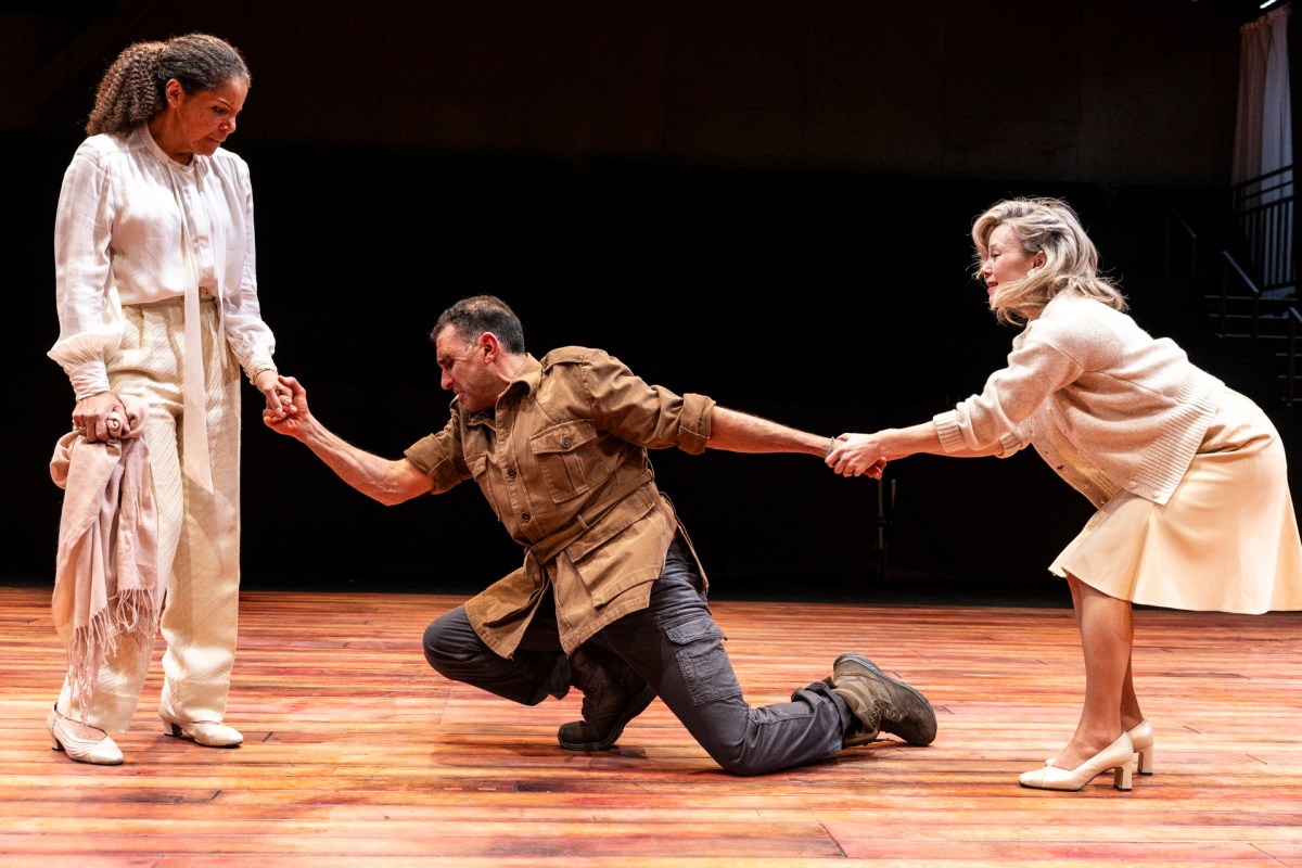 A man in brown is kneeling between two women in white.