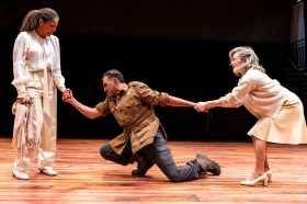 A man in brown is kneeling between two women in white.