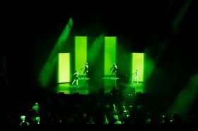 Four dancers stand poised on a gree-lit stage as a DJ plays at stage front. An audience looks on. This is a sequence from 'Cortex' by the Kianí del Valle Performance Group, which has been programmed alongside cutting edge sound works and DJs at Melbourne Town Hall for Now or Never.