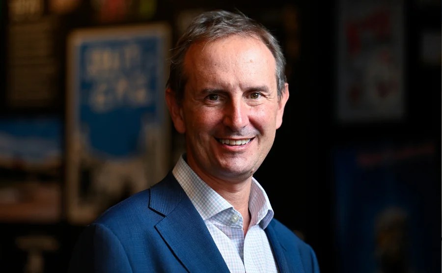 A middle aged man in a white open collar shirt and blue jacket smiles at the camera. Alex Budd of Canberra Theatre Centre.