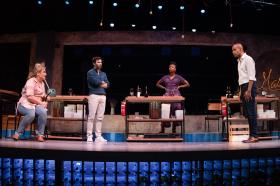 Two men and two women stand beside small work tables.
