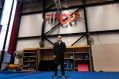 A grey-haired and grey beared fair-skinned man wearing a long black coat and dark framed glasses, stands in a circus training facility. The word 'Circus' in colourful lettering hangs on the wall behind him.