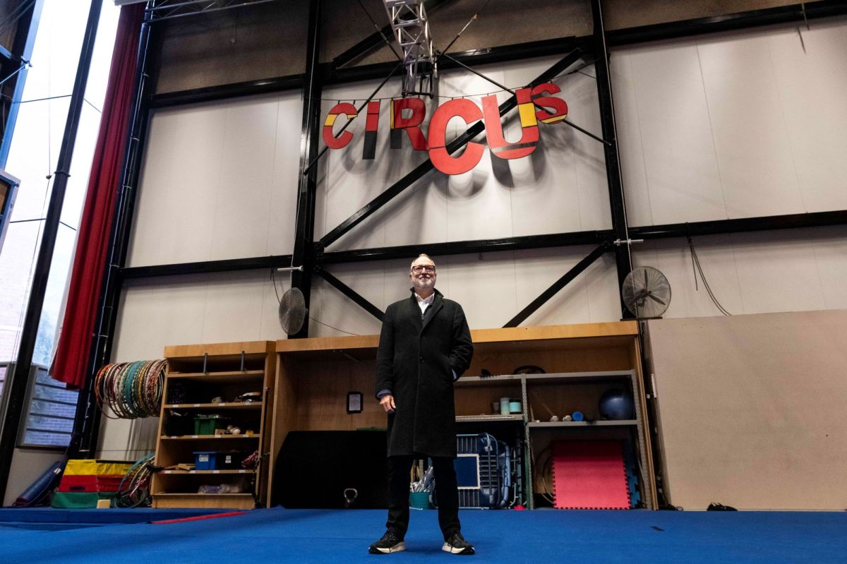 A grey-haired and grey beared fair-skinned man wearing a long black coat and dark framed glasses, stands in a circus training facility. The word 'Circus' in colourful lettering hangs on the wall behind him.