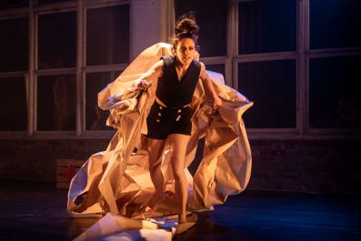 A fair-skinned woman, wearing black shorts and a black waistcoat, stands in a constricting shroud of paper (perhaps representing paperwork) on a bare, industrial-looking stage. This is Andi Snelling in her solo show 'Happy-Go-Wrong' at fortyfivedownstairs.