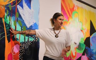 A woman wearing a large necklace and white tshirt placing her hand on a large artwork of colourful abstract shapes.