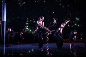 Dancers in black costume. The stage and lighting are also black.