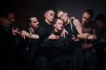 contemporary dance: a dark stage with the faces and upper bodies of five dancers costumed in black. Their faces and arms standout against the black as they hold each other in a communal embrace.