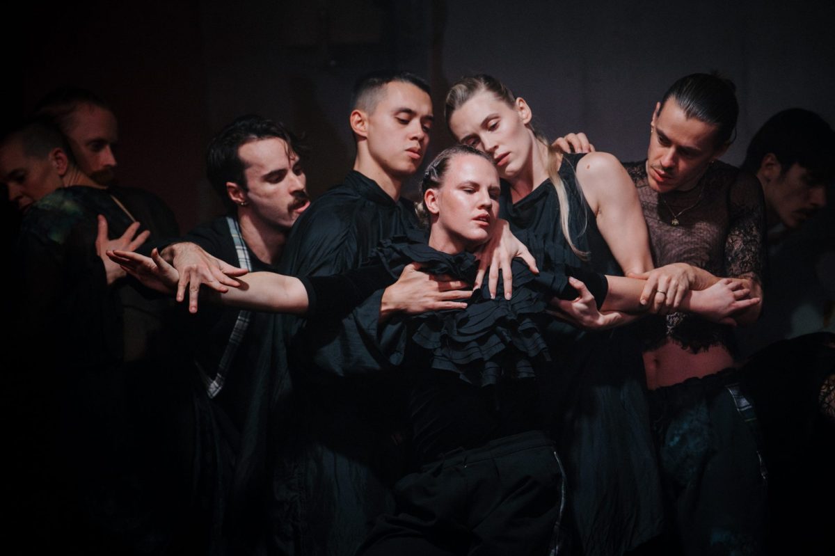 contemporary dance: a dark stage with the faces and upper bodies of five dancers costumed in black. Their faces and arms standout against the black as they hold each other in a communal embrace.