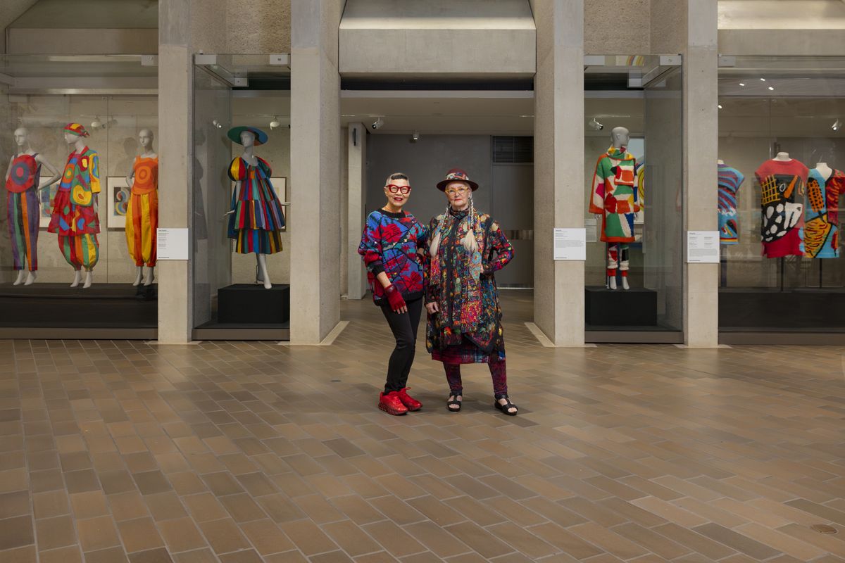 Jenny Kee (an older Asian-Australian woman wearing tight black jeans matched with red shoes, gloves and glasses which echo the vivid designs of her boldly colourful jumper) and Linda Jackson (an older fair-skinned woman with her hair in braids and wearing sandals, red leggings and a vibrantly coloured long-sleeved, knee length dress and matching hat) pose with pieces from the 'Know My Name: Kee, Jackson and Delaunay' exhibition at the National Gallery of Australia, Kamberri/Canberra, 2025.
