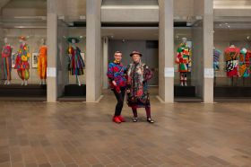 Jenny Kee (an older Asian-Australian woman wearing tight black jeans matched with red shoes, gloves and glasses which echo the vivid designs of her boldly colourful jumper) and Linda Jackson (an older fair-skinned woman with her hair in braids and wearing sandals, red leggings and a vibrantly coloured long-sleeved, knee length dress and matching hat) pose with pieces from the 'Know My Name: Kee, Jackson and Delaunay' exhibition at the National Gallery of Australia, Kamberri/Canberra, 2025.