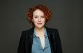 A fair-skinned woman with curly red hair looks quizzicaly at the camera, one eyebrow slightly raised and a playful expression on her face. She wears a formal black jacket over a light blue blouse, which is unbuttoned, giving her a somewhat devil-may-care attitude. This is Kerri Glasscock, the outgoing Director and CEO of Sydney Fringe Festival.